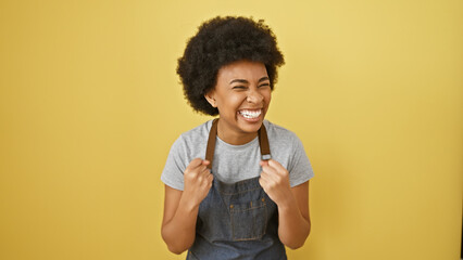 Cheerful african american woman with curly hair wearing an apron smiles against a yellow wall.