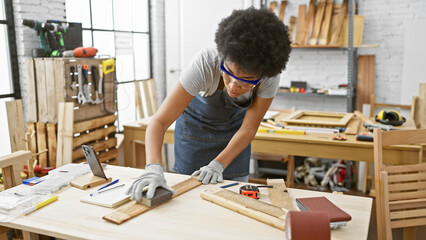 Focused black woman sanding wood in a well-equipped carpentry workshop, wearing protective goggles.