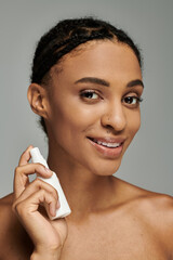 Young African American woman in strapless top smiles while holding a cosmetic product on grey...