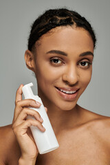 Young African American beauty in strapless top delicately holds a bottle of cleanser on a grey...