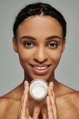 African American woman holds cream jar in front of face against grey background, taking skincare...