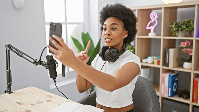 African american woman recording in a music studio, holding a smartphone, with headphones around her neck.