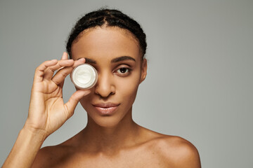 Young African American woman in strapless top holds cream jar in front of face, skin care concept,...