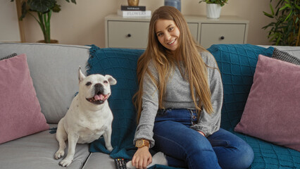 A young blonde woman sits on a sofa in a cozy living room with her cheerful canine pet beside her.