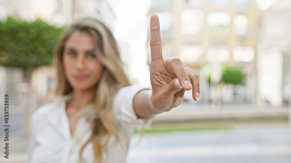 Wall mural Cool, young blonde woman exudes relaxed, yet serious energy, standing outdoors in sunny, urban street, resolutely saying no with her finger while maintaining eye contact with camera