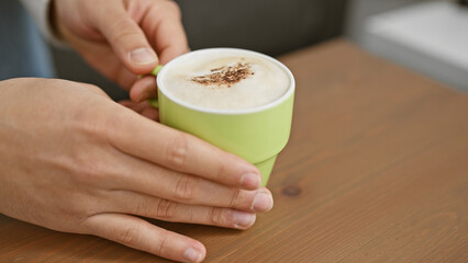 Close-up of a man's hands holding a green mug of cappuccino on a wooden table indoors. - Powered by Adobe