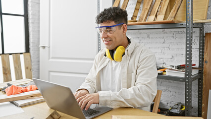 Smiling man with headphones using laptop in a workshop full of carpentry tools.