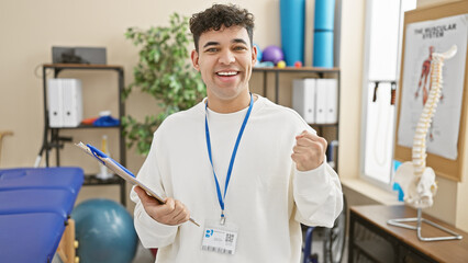 A cheerful young man with a badge and clipboard celebrates success in a bright physiotherapy clinic.