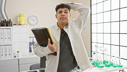 A pensive young man holding a folder stands in a laboratory, reflecting thoughtfulness and...