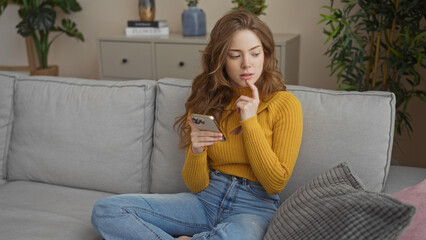 A young, attractive blonde woman seated in a home living room, engaging with her phone while...