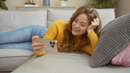 A beautiful young blonde woman lies on a couch in the living room, smiling at her smartphone in a cozy home setting.