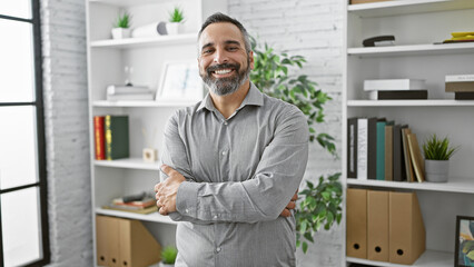 Confident mature hispanic man with grey beard standing arms crossed in a modern office setting,...