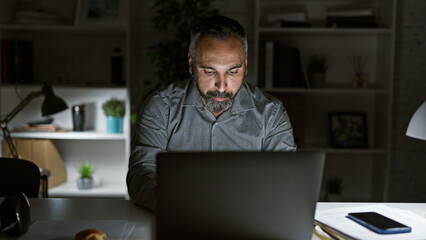 Hispanic senior man with grey beard working late night in a dark office interior, focused on laptop...