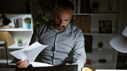 A mature hispanic man with grey beard reviews documents at night in a dark office. © Krakenimages.com