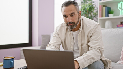 Mature hispanic man with grey beard using laptop in a modern living room.