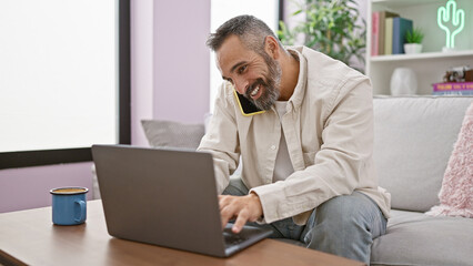 Smiling mature man with grey beard multitasking with phone and laptop in a cozy living room.