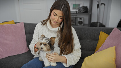 A young hispanic woman cuddles a biewer yorkshire terrier in a cozy living room, conveying domestic...