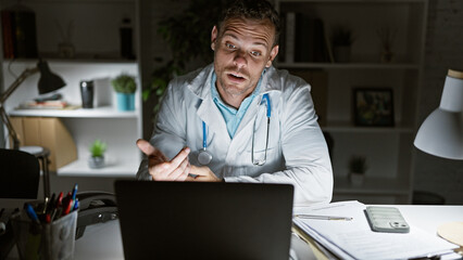 A hispanic male doctor in a white coat gestures during a video call in a dimly lit clinic office at night