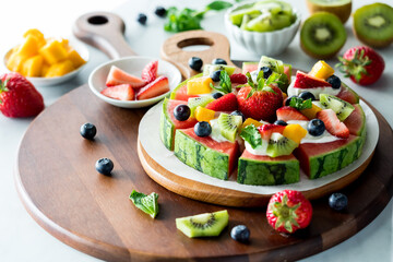 A watermelon pizza on a wooden platter surrounded by fresh fruit toppings.