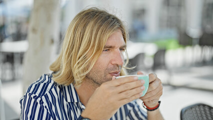 A pensive blond man enjoys coffee outdoors on a sunny urban terrace, depicting leisure in casual...