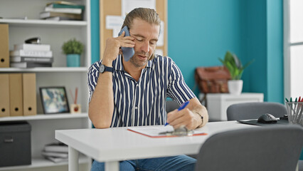 A businessman multitasking in an office, talking on the phone while writing notes, surrounded by...