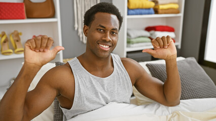 Smiling african american man sitting in bedroom pointing at himself with thumbs, casual, comfort,...