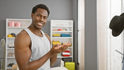 A smiling young black man in a sleeveless shirt presenting with hands in a well-organized bedroom...