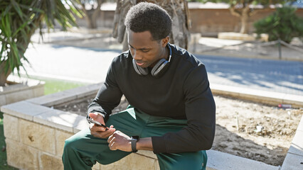 A young african american man with headphones and a smartwatch checks his phone in a sunny outdoor...