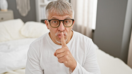 A mature man with glasses gestures silence in a bedroom setting, expressing a concept of privacy...