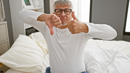 A middle-aged man with grey hair gives a thumbs down while sitting on a white bed in a bedroom...