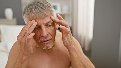 A shirtless, gray-haired man appears stressed or in discomfort, sitting on a bed indoors, evoking...