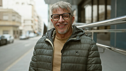 A smiling middle-aged man with grey hair poses casually outdoors on a city street, exuding...