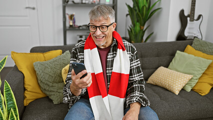 A cheerful, mature man interacts with a smartphone on a cozy sofa, surrounded by stylish home decor. - Powered by Adobe
