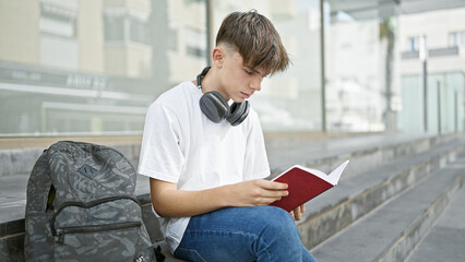 A young caucasian male teenager with headphones around his neck reads a book on urban steps, with a...