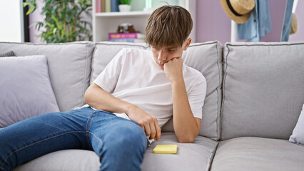 Blond teenager boy reflecting alone on a gray sofa indoors, conveying a thoughtful mood in a casual home setting.