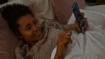 A relaxed woman with curly hair enjoys leisure time browsing on her smartphone in her cozy bedroom.