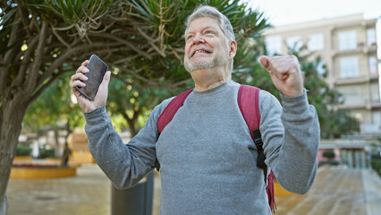 Smiling senior man with smartphone and backpack celebrating success outdoors in the city.