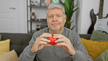 A grey-haired man holds a red heart in a cozy living room, symbolizing love and health awareness.