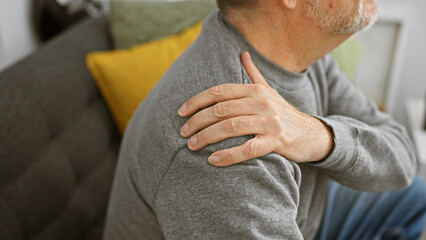 A mature grey-haired man experiences shoulder pain while sitting in a living room.