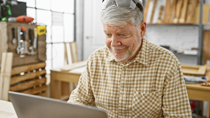 A senior man with grey hair smiles while using a laptop in a carpentry workshop.