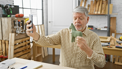 Handsome senior man taking a selfie with wooden horse in a creative carpentry workshop