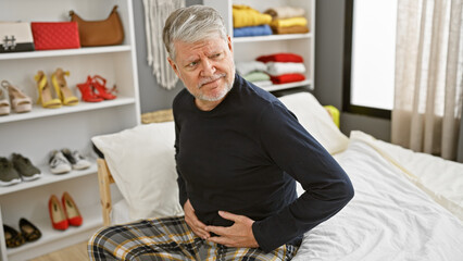 Senior man with gray hair experiencing stomach pain while sitting on a bed in a well-lit bedroom