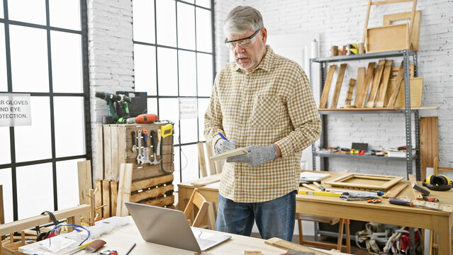 Middle-aged man writing in notebook in a woodworking workshop with tools and laptop. - Powered by Adobe