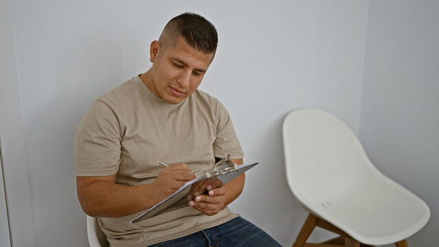 Serious latin guy, relaxed yet concentrated, sits in chair taking notes. indoor portrait of young hispanic man writing job interview checklist on clipboard in waiting room background.