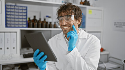 Handsome hispanic man with beard and green eyes smiling in a laboratory while holding a tablet,...