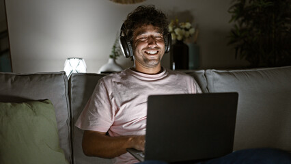 A smiling young hispanic man relaxes at home, enjoying music on headphones with a laptop at night.