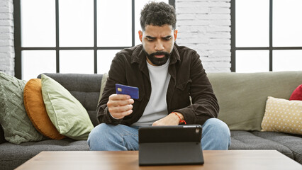 Hispanic man using tablet and holding credit card indoors at home, sitting on couch with beard and casual attire.