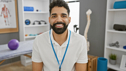 A handsome young man with a beard smiles warmly, wearing a lanyard in a well-equipped physical...