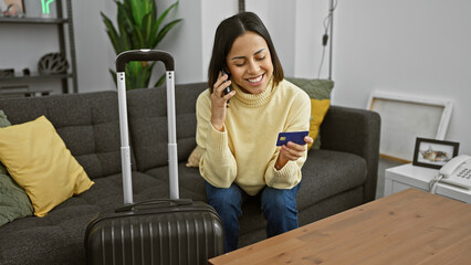 A young hispanic woman smiles while holding a credit card and talking on a smartphone, sitting indoors with a suitcase by her side.