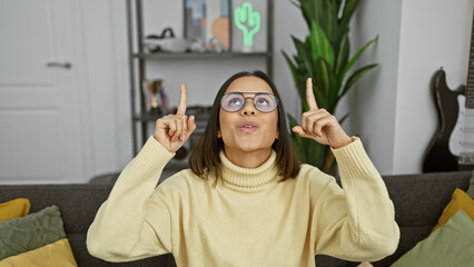 Fototapeta premium Hispanic woman pointing upwards with a playful expression indoors, exuding youth and positivity in a cozy living room setting.